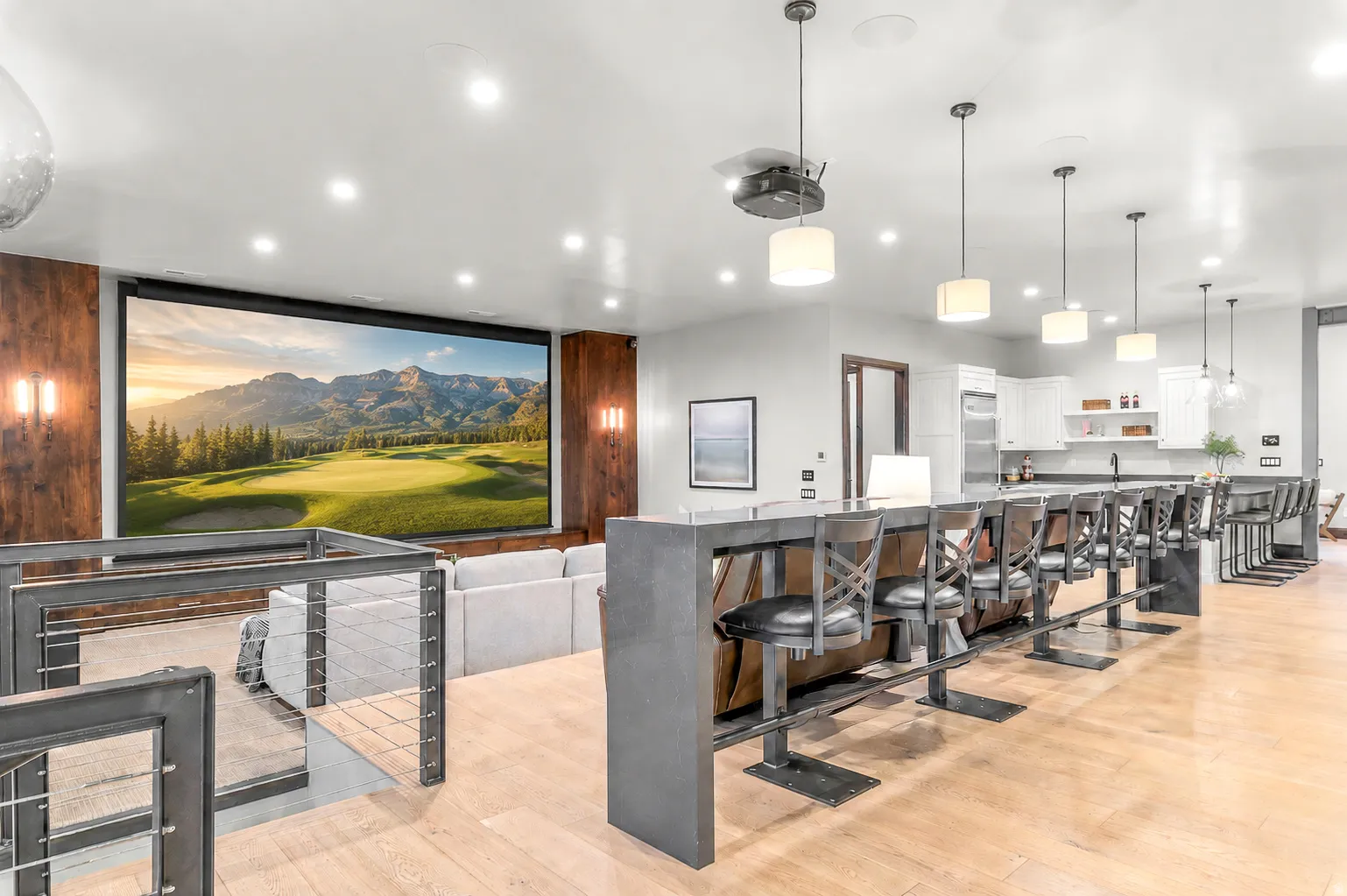 Expansive entertainment space featuring a large projection screen, built-in bar seating, wood-finish flooring, recessed lighting, and a full kitchen with white cabinetry