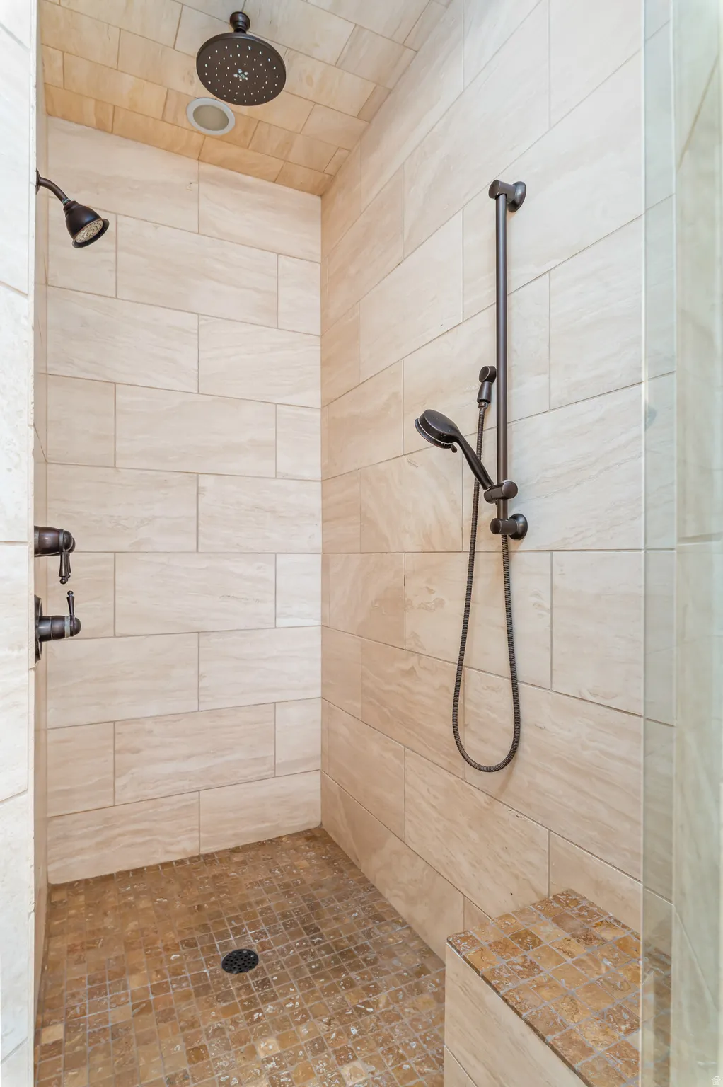 Walk-in shower with neutral-toned wall tiling, mosaic tile floor, built-in bench, and dual shower heads