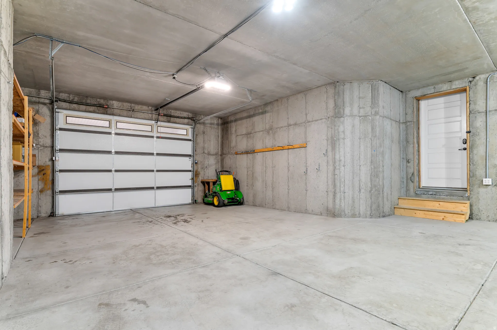 Concrete flooring, exposed concrete walls, white overhead garage door with windows, white side entry door, and timber shelving