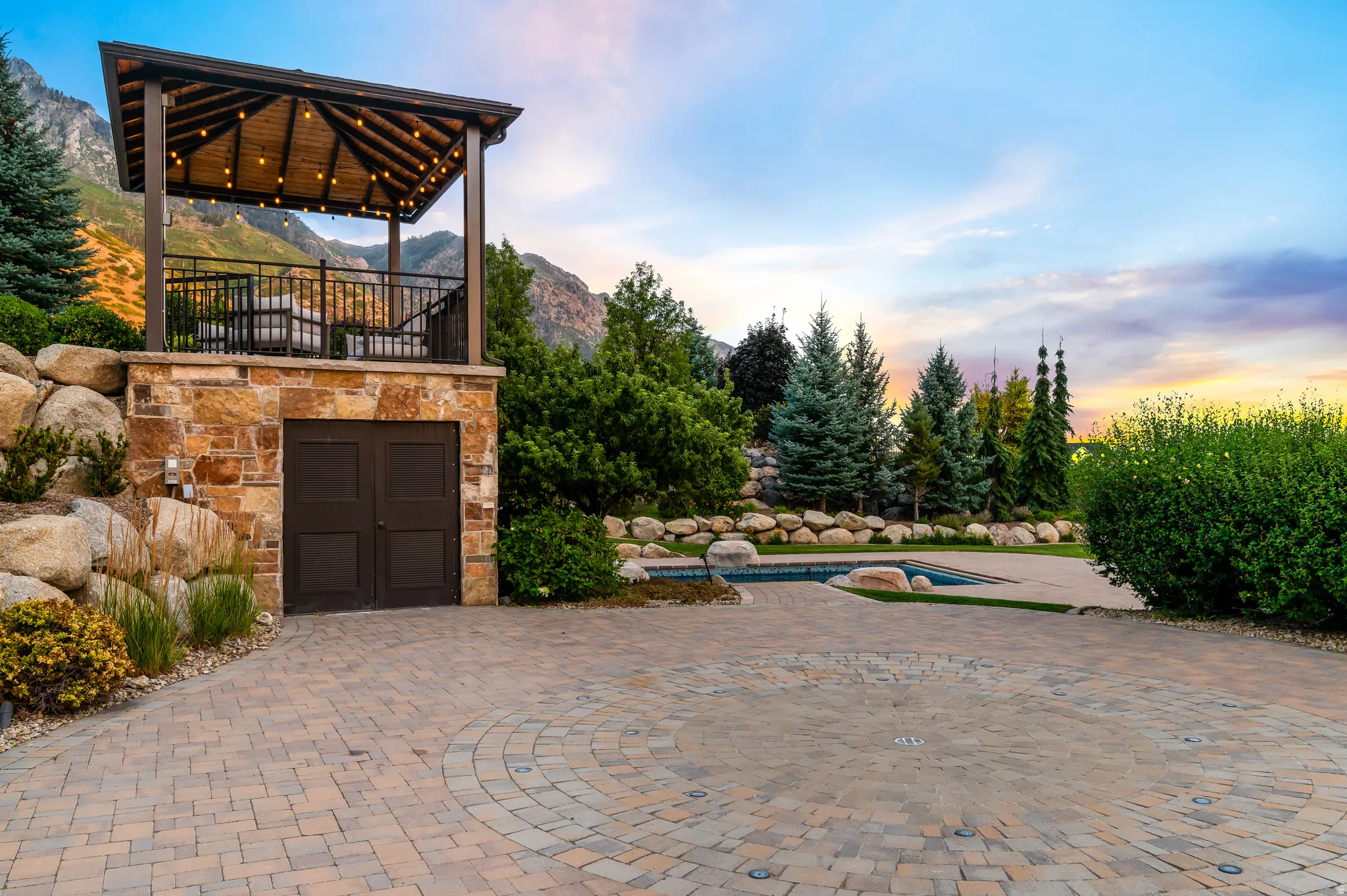 Paved driveway with a circular pattern, stone-clad structure with a covered balcony, integrated lighting, and a visible pool area