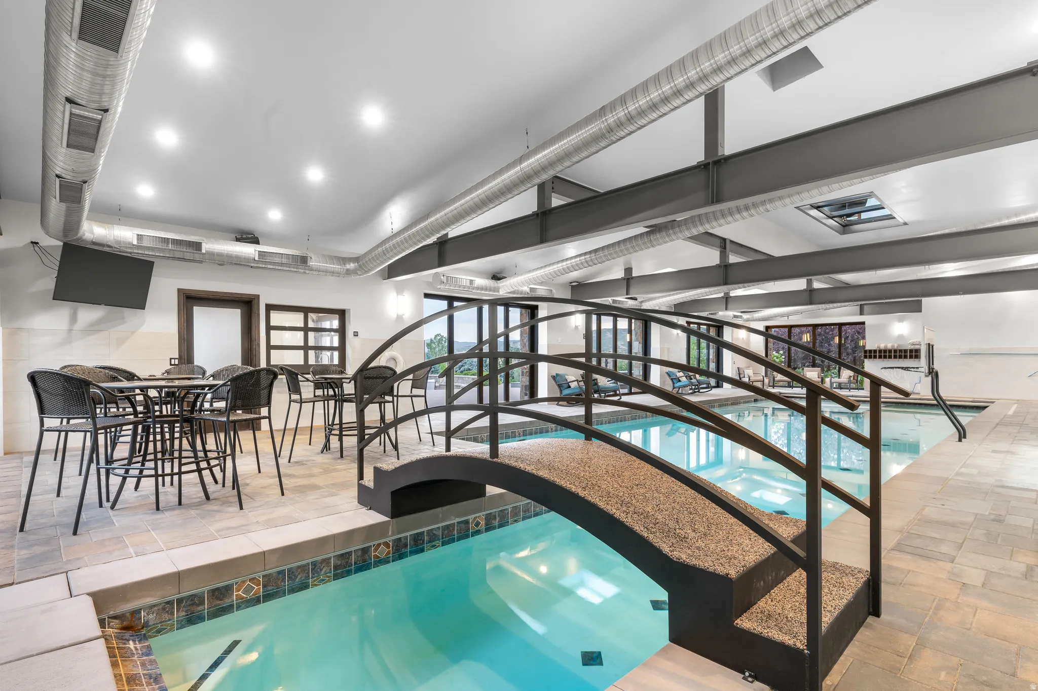 Indoor swimming pool with an arched footbridge, exposed structural beams, ceiling-mounted recessed lighting, and large glass doors
