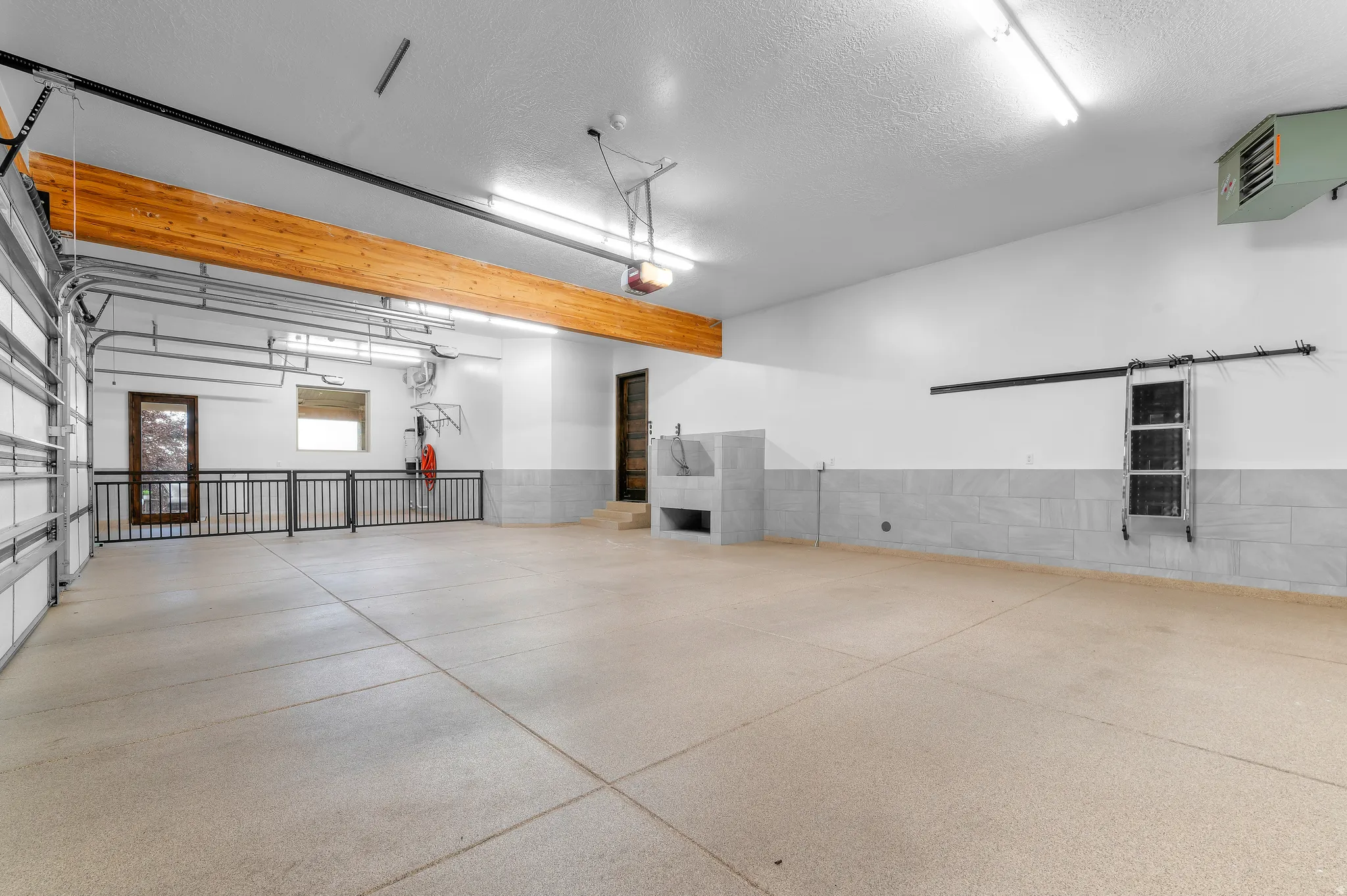 Spacious garage with light-colored flooring, exposed wood beam, wall-mounted utility sink, and wall-mounted heating unit