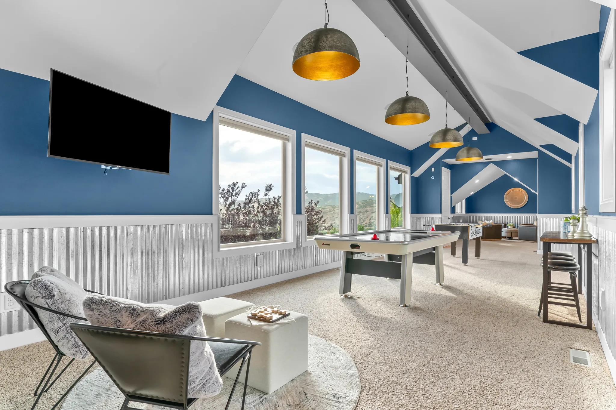 Vaulted ceiling with exposed beams, multiple windows, corrugated metal wainscoting, wall-mounted television, and pendant lighting