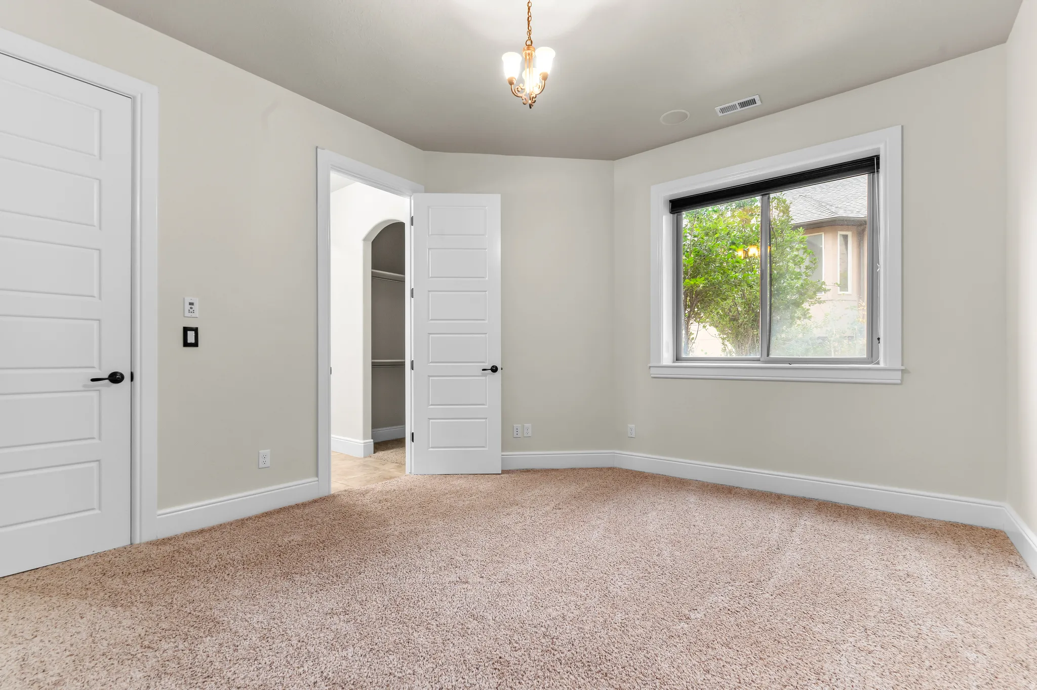 Plush carpet flooring, neutral painted walls, a built-in closet with shelving, a white-framed window, and a decorative chandelier