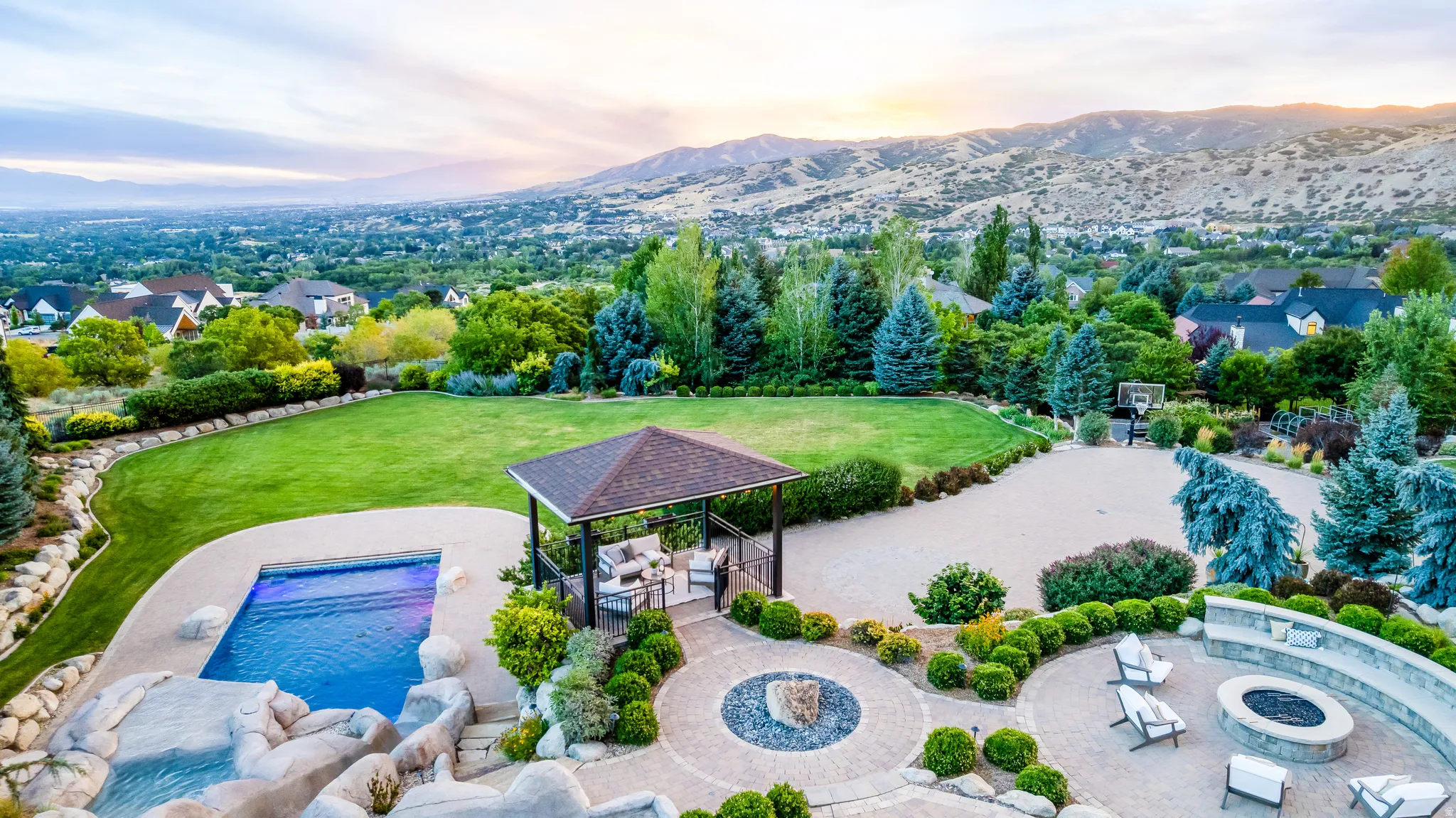 Expansive rear grounds featuring a swimming pool with rock surround, paved patio with a built-in fire pit, covered gazebo, and a large lawn area