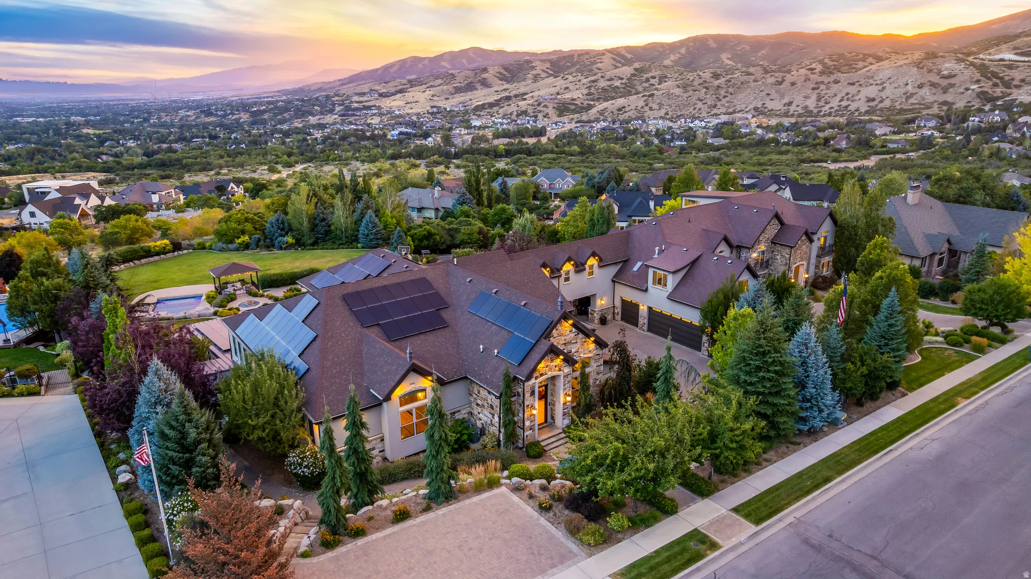 Stone and stucco exterior, multiple roof-mounted solar panels, integrated three-car garage, expansive green lawn, and a large inground pool