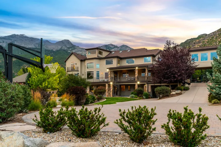Multi-level stucco exterior with stone accents, featuring extensive landscaping, a paved driveway, and a prominent mountain backdrop