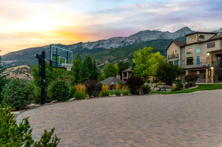 Paver driveway with a basketball hoop, multi-level exterior, stone accents, and mature landscaping