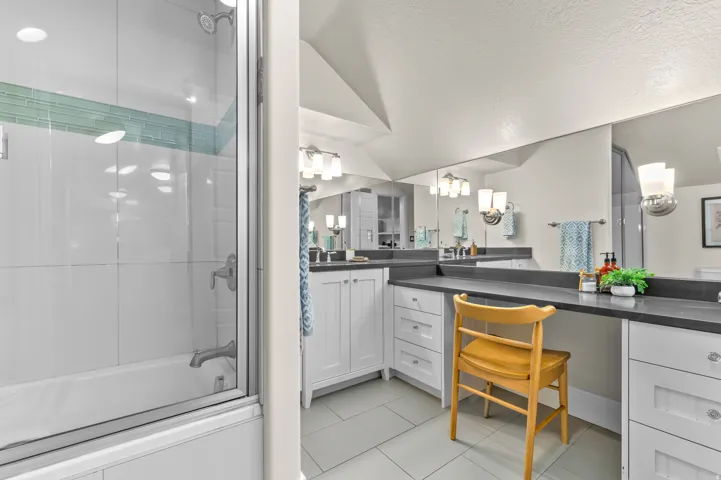 White cabinetry with brushed nickel hardware, expansive vanity with dark countertops, built-in vanity station, recessed lighting, and a glass-enclosed shower/tub combination with subway tile surround and decorative accent tiling