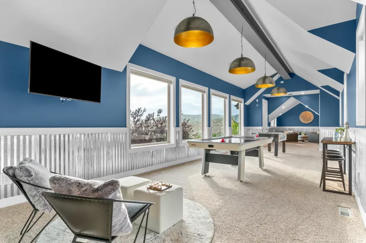 Vaulted ceiling with exposed beams, multiple windows, corrugated metal wainscoting, wall-mounted television, and pendant lighting