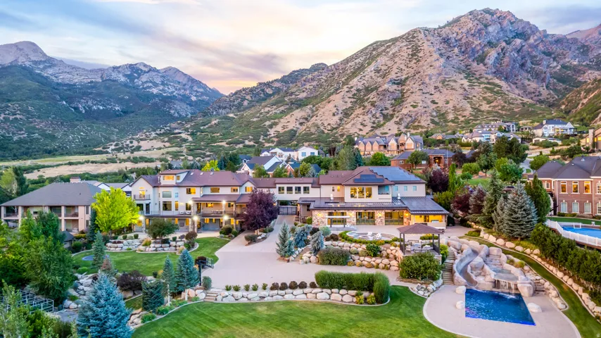 Expansive property with a large main residence, terraced landscaping, a built-in outdoor pool with a rock water feature, and a paved driveway, all framed by distant mountains