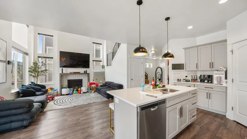 Open concept living area featuring, a kitchen island with sink and integrated dishwasher, light gray cabinetry,  and a fireplace with a white brick surround