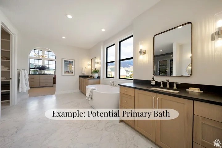 Freestanding soaking tub, dual vanities with dark countertops, wood-finish cabinetry, marble-look flooring, and large windows