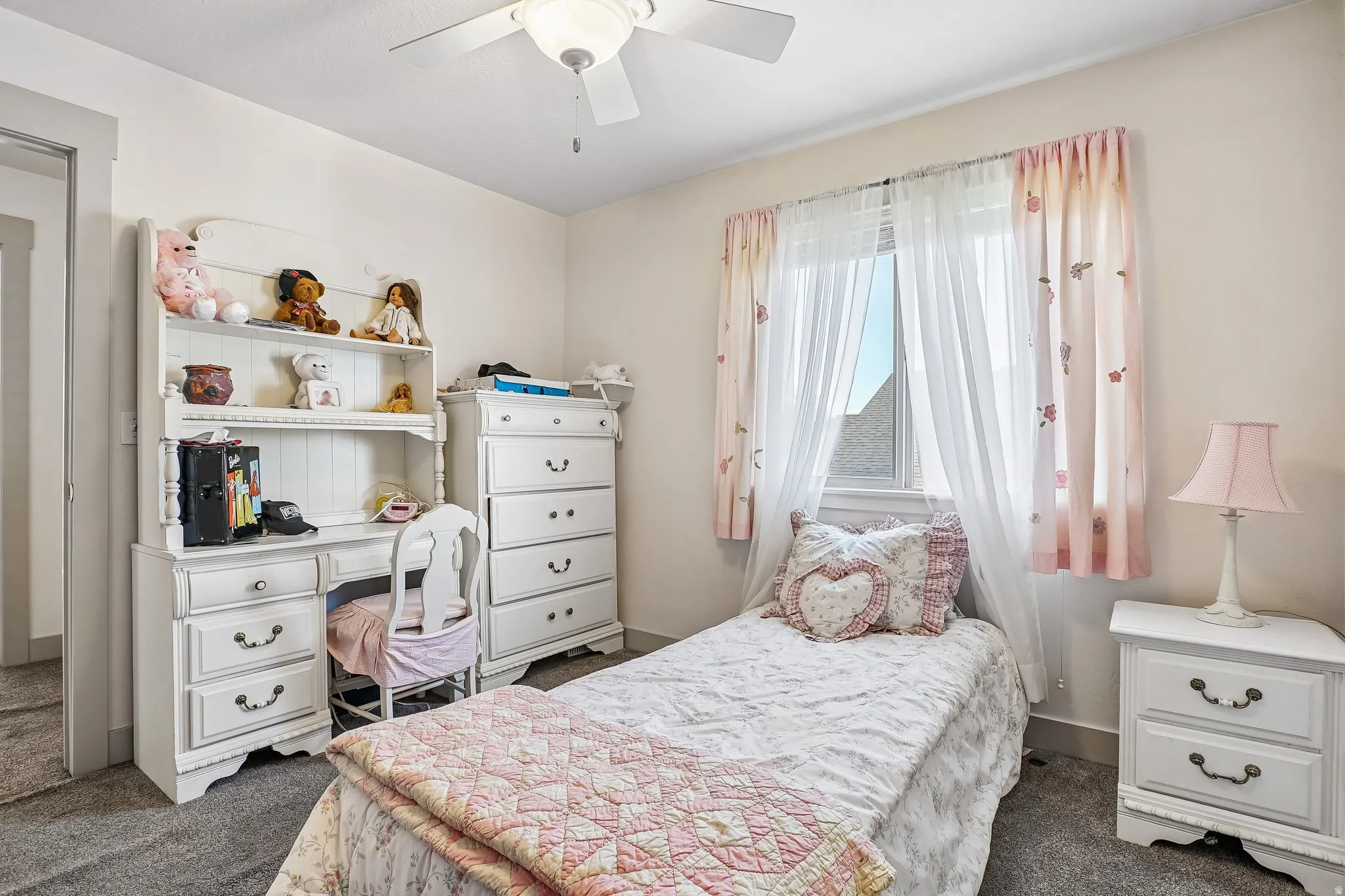 Carpeted flooring, ceiling fan with integrated lighting, built-in desk unit with shelving, window with natural light, and a white nightstand