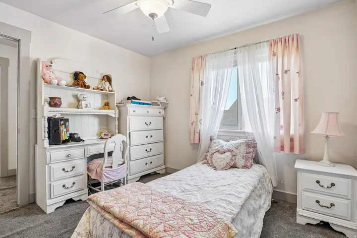 Carpeted flooring, ceiling fan with integrated lighting, built-in desk unit with shelving, window with natural light, and a white nightstand