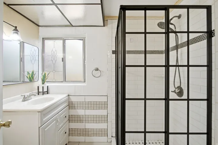 White vanity with integrated sink, black grid-patterned shower enclosure, subway tile shower surround, and a leaded glass window