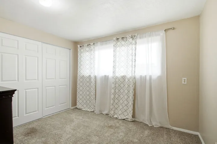 Plush carpet flooring, white paneled bifold closet doors, and a window with sheer and patterned curtains