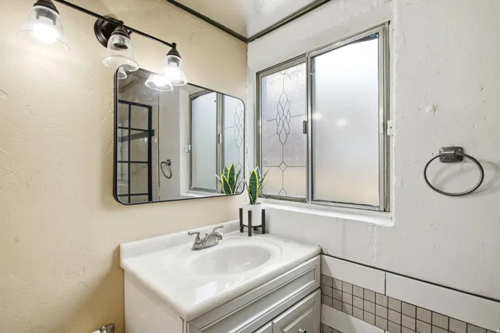 Vanity with integrated sink, rectangular mirror, three-light vanity fixture, tiled wainscoting, and frosted window