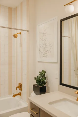 Bathtub with vertical striped tile surround, gold-tone showerhead and faucet, under-mount sink, and dark wood-finish vanity with gold-tone hardware