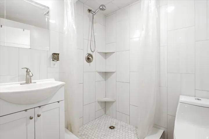 Compact bathroom featuring a white vanity with cabinet storage, a wall-mounted mirror, a shower with built-in corner shelving, white wall tile, and a white hexagon tile shower floor
