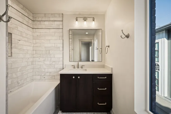 Tub with subway tile surround, dark wood-finish vanity with countertop, framed mirror, and brushed nickel hardware
