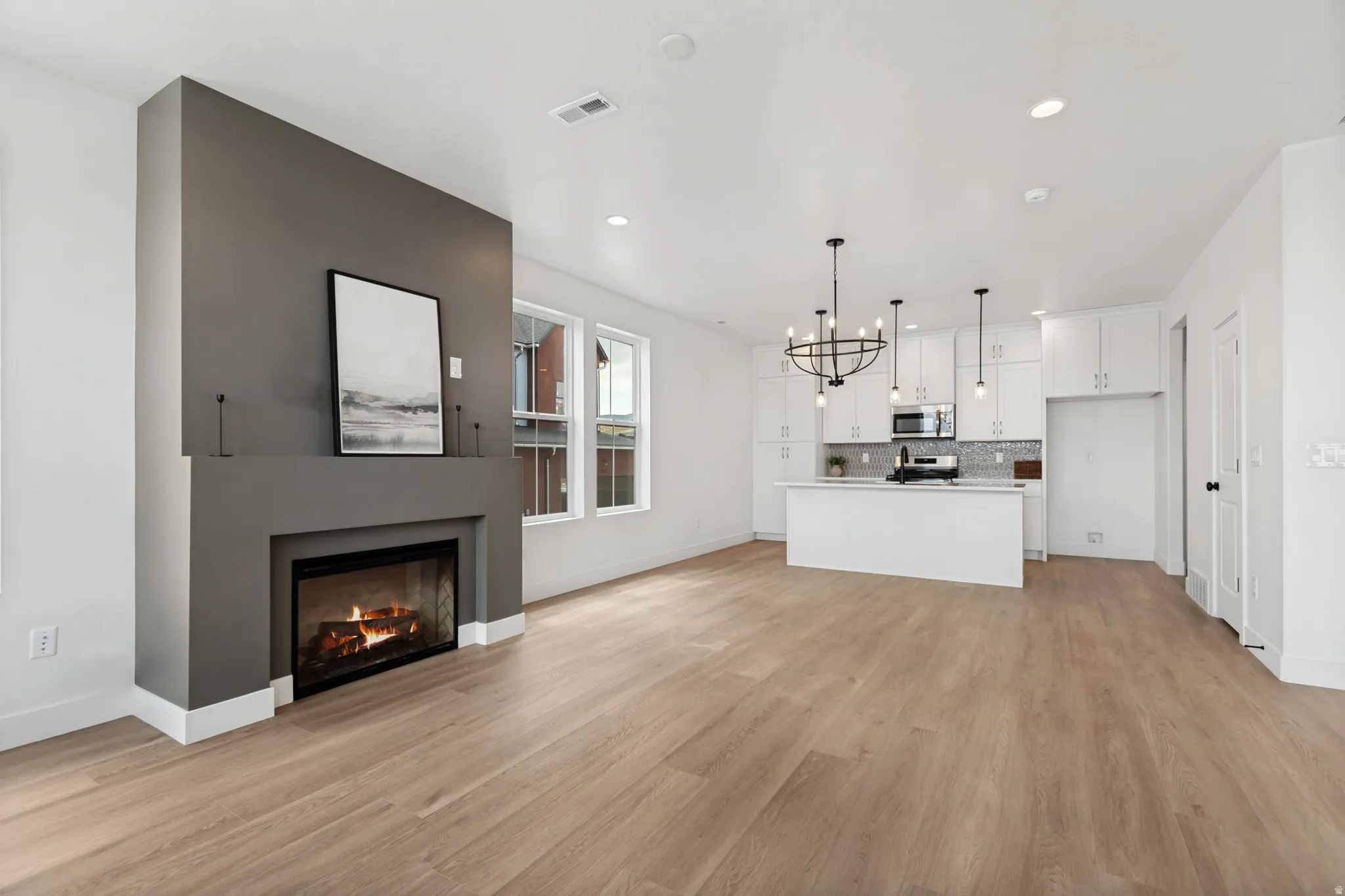 Open-concept living space with wood-finish flooring, a contemporary fireplace, and a kitchen featuring white cabinetry, an island, and pendant lighting