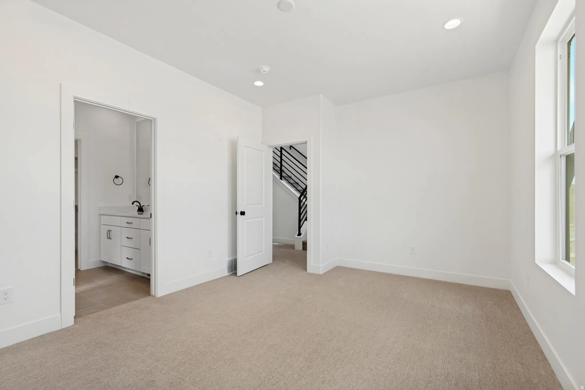 Neutral tone wall paint, recessed ceiling lighting, light-colored carpeting, white trim window, and a white vanity with a black faucet