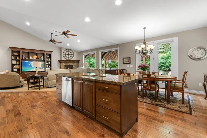 Open-concept kitchen and dining area featuring a large center island with wood cabinetry and stone countertops, stainless steel appliances, hardwood flooring throughout, recessed and pendant lighting, and abundant natural light from multiple windows and g