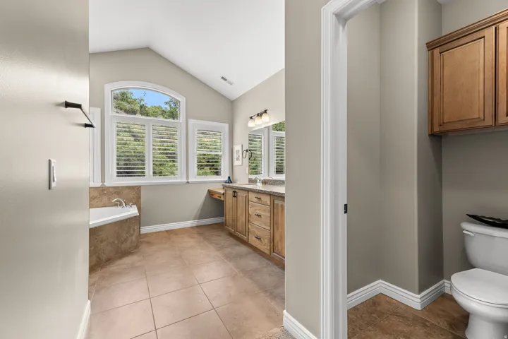 Vaulted ceiling with an arched window, plantation shutters, built-in soaking tub with tile surround, double vanity with stone countertop, and tile flooring