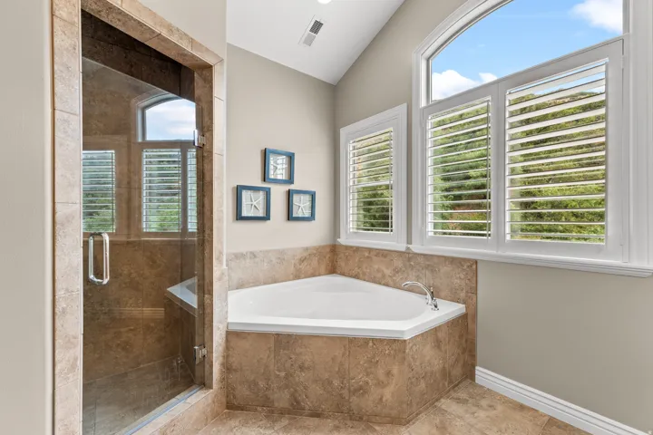 Tile-surrounded corner soaking tub with deck-mounted fixtures, separate glass-enclosed shower with tile surround, and arched window with white plantation shutters