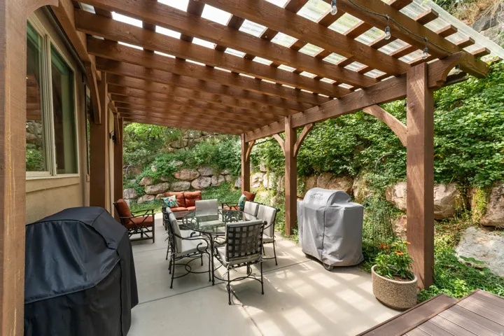 Wood pergola with a translucent roof, featuring string lighting, a concrete patio, and a stone retaining wall