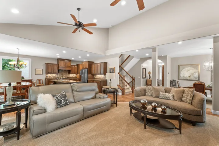 Vaulted ceiling with recessed lighting and ceiling fans, open concept living area, staircase with wood banister, kitchen with wood cabinetry and stone countertops
