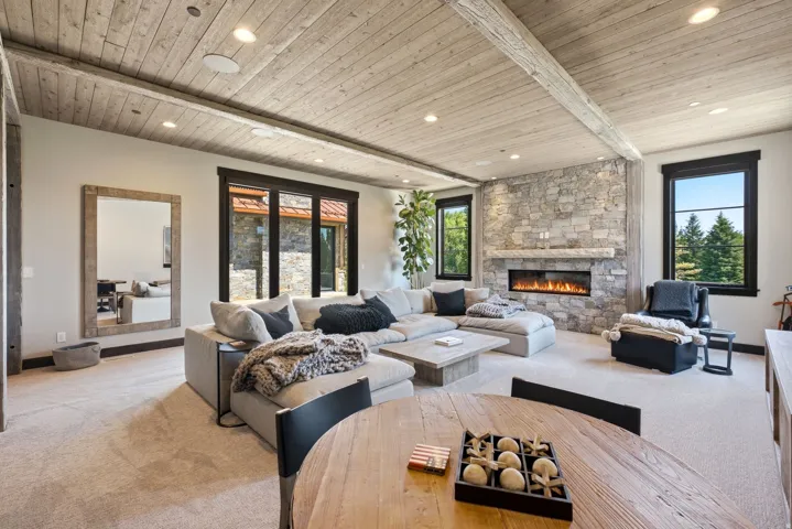 Coffered ceiling with exposed wood beams, stone-clad fireplace with a linear insert, recessed lighting, and large windows with dark trim