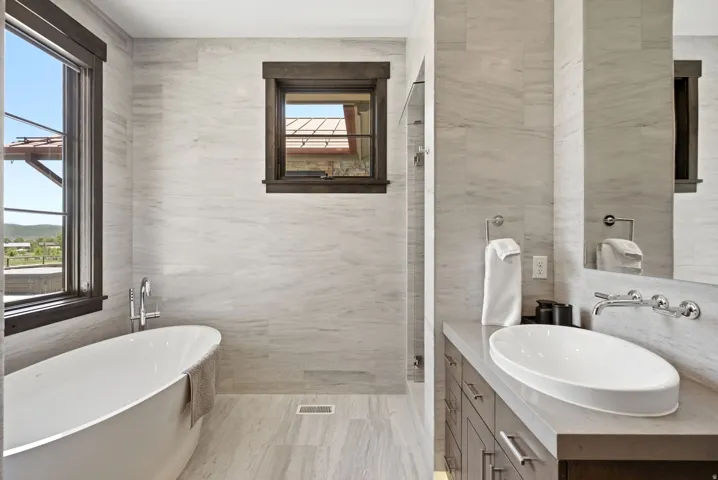 Freestanding soaking tub, floor-to-ceiling tile, dark wood-framed windows, vanity with vessel sink, and wall-mounted faucet