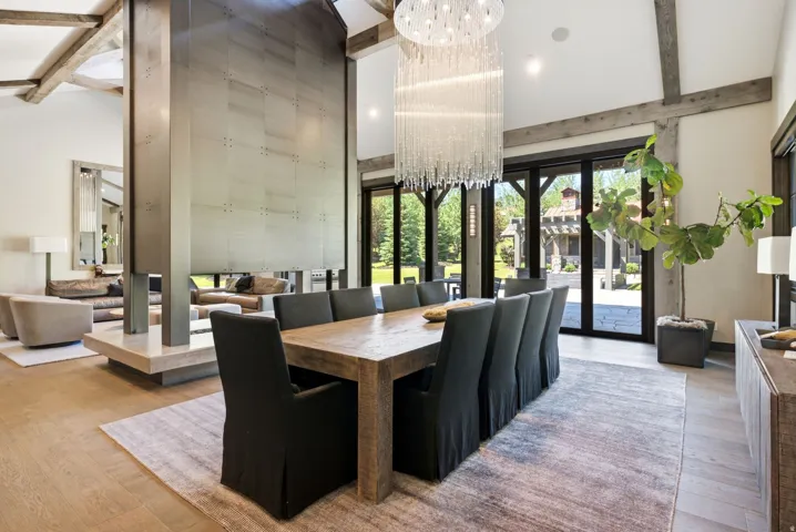 Open-concept interior featuring wood-finish flooring, exposed wood beams, a towering fireplace, a large modern chandelier, and extensive glass doors