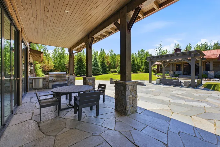 Expansive stone patio featuring a covered outdoor kitchen with a built-in grill and sink, a large wood pergola, and abundant surrounding greenery