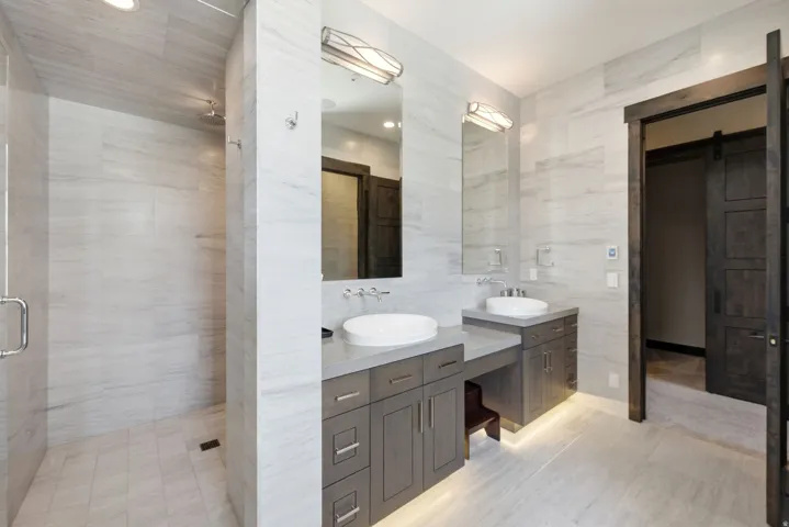 Walk-in shower with tile surround, dual floating vanities with vessel sinks, integrated lighting under vanities, and a dark wood-finish sliding barn door