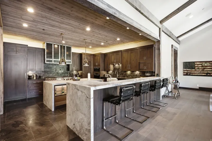 Wood-paneled ceiling with recessed lighting, stone tile flooring, waterfall island with integrated beverage cooler, dark wood cabinetry, and exposed wood beams