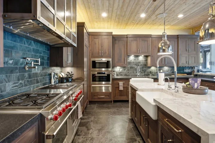Gourmet kitchen featuring a wood plank ceiling, stone tile flooring, an oversized island with a farmhouse sink, stainless steel appliances, and dark wood cabinetry