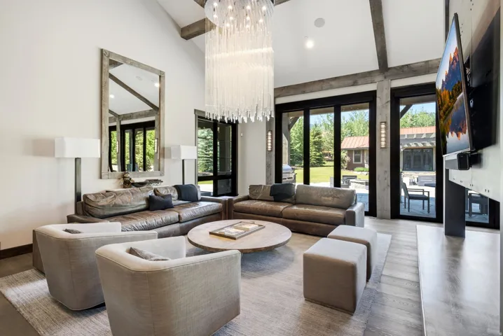 Spacious living area featuring a vaulted ceiling with exposed beams, wood-finish flooring, a large decorative chandelier, and expansive glass doors opening to the exterior