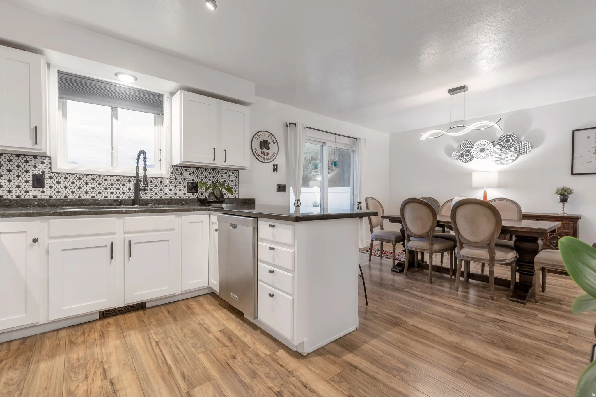 White cabinetry with dark hardware, dark countertops, patterned tile backsplash, stainless steel dishwasher, and wood-finish flooring