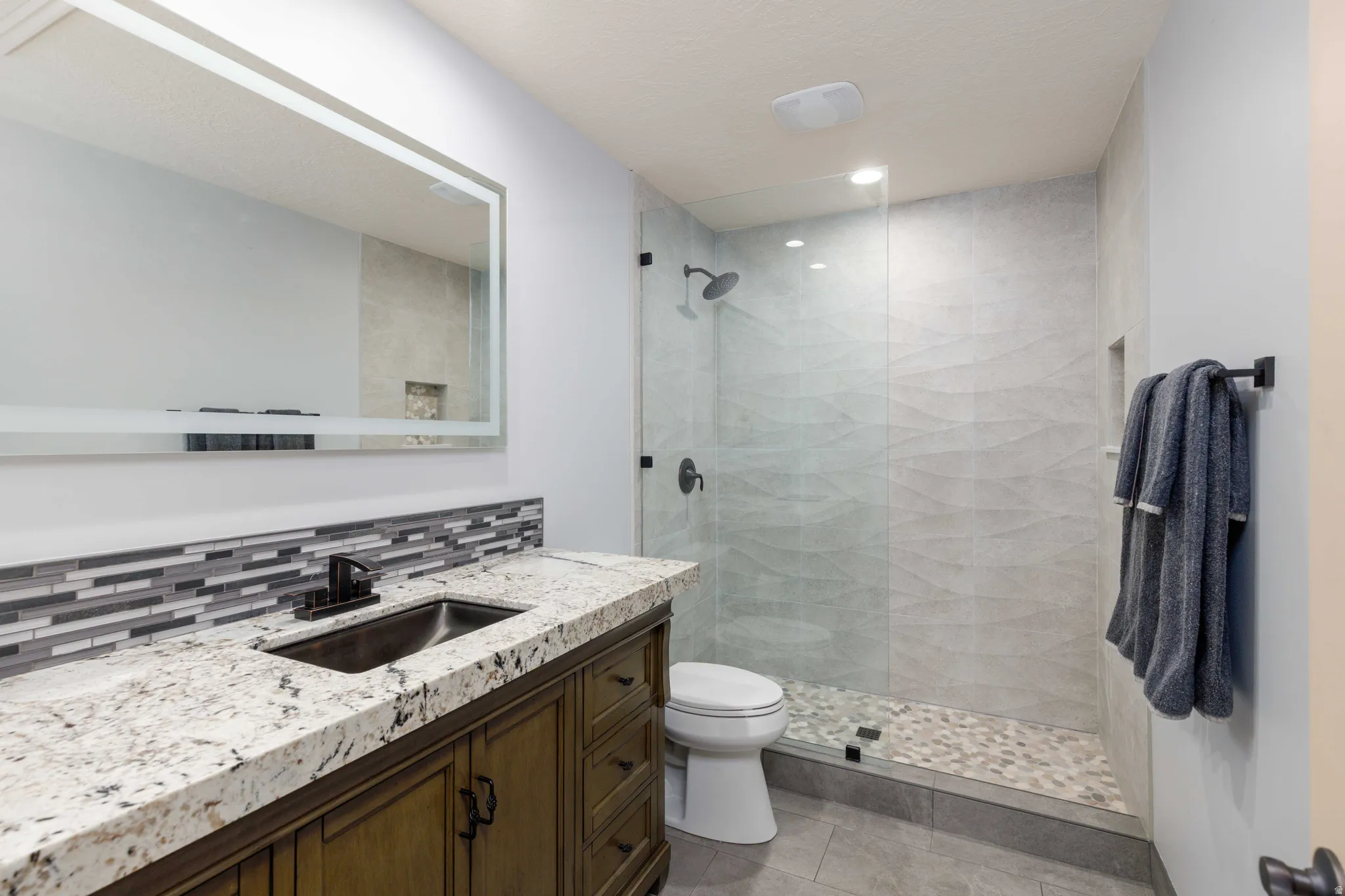 Granite countertop vanity with undermount sink, mosaic tile backsplash, and frameless glass shower enclosure featuring a pebble floor