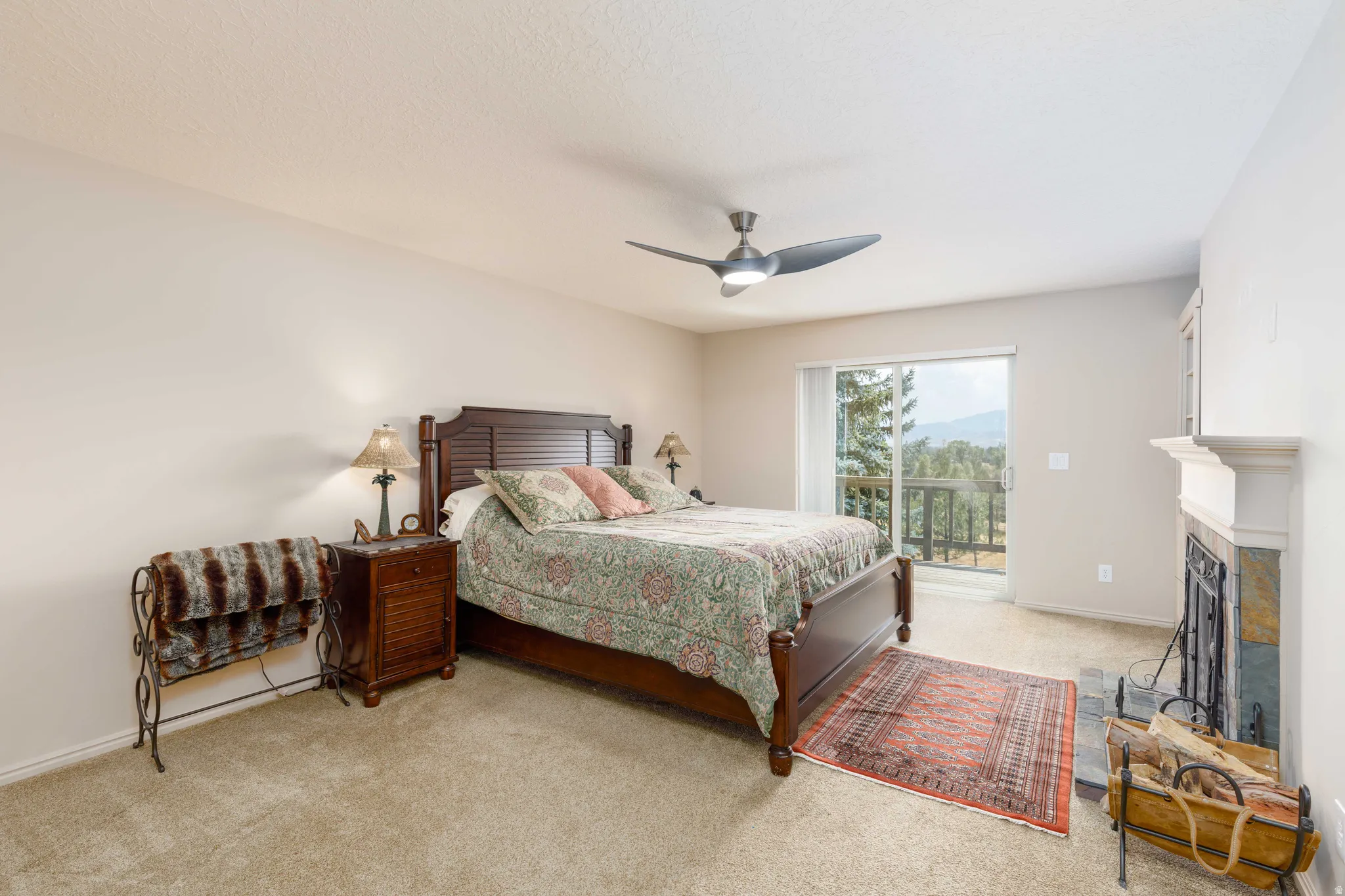 Carpeted bedroom featuring a ceiling fan, a fireplace with a stone surround and white mantel, and a sliding glass door opening to an exterior deck