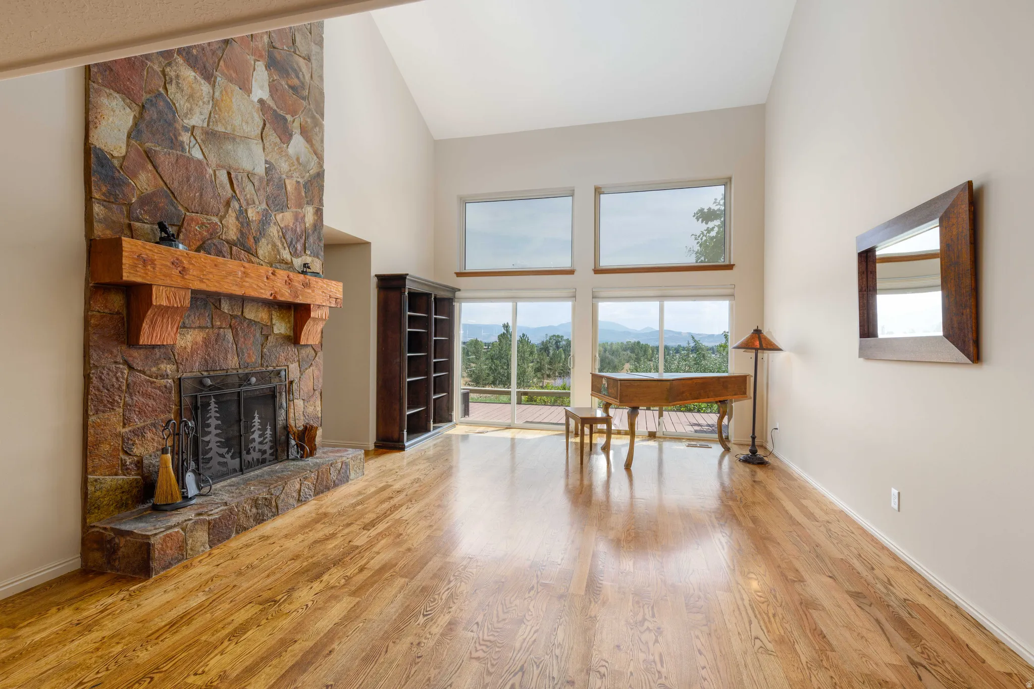 Vaulted ceiling, stone fireplace with wood mantel, wood-finish flooring, built-in shelving, and large windows with outdoor scenery