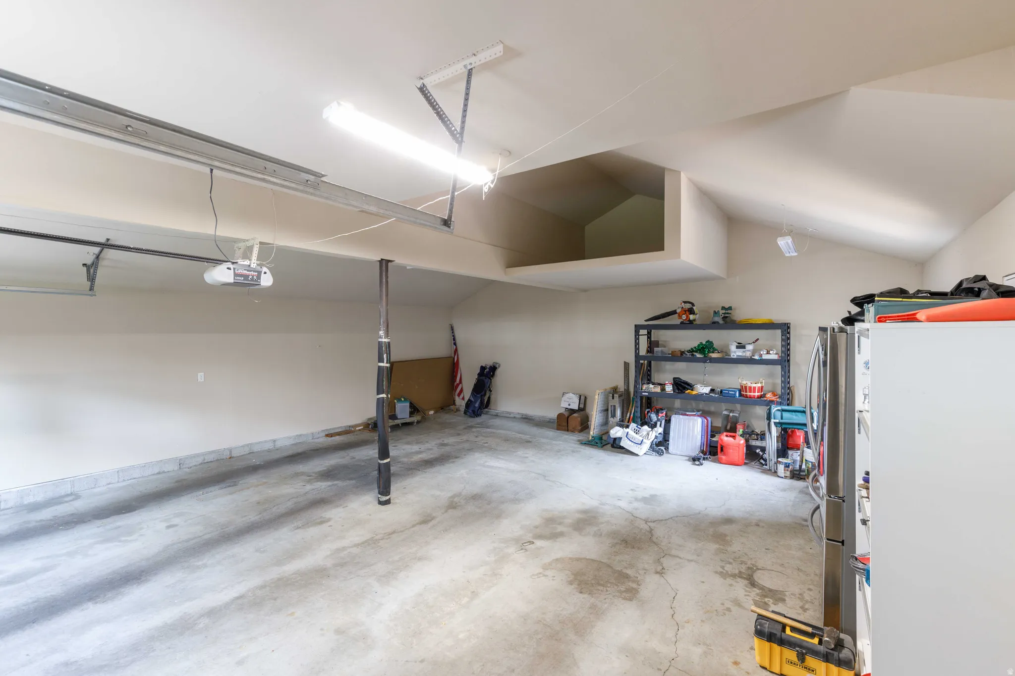 Spacious garage with concrete flooring, overhead fluorescent lighting, and an integrated storage loft