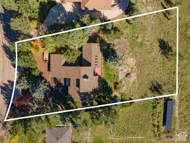 Aerial perspective of a sprawling lot featuring mature trees, natural landscape, a roof with integrated skylights, and a ground-mounted solar array