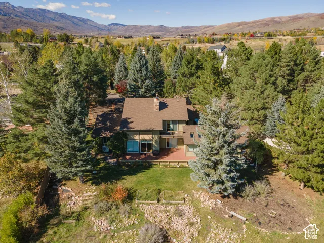 Multi-level exterior featuring a shingled roof, chimney, attached rear deck, and sliding glass doors, set within a landscaped lot with mature trees and mountain backdrop