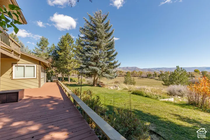 Expansive elevated wood deck featuring horizontal railing, mature pine trees, manicured lawn, and mountainous horizon