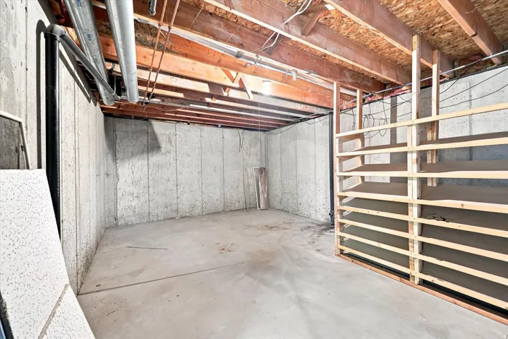 Unfinished basement with exposed wood joists, concrete foundation walls, concrete flooring, and built-in wood shelving