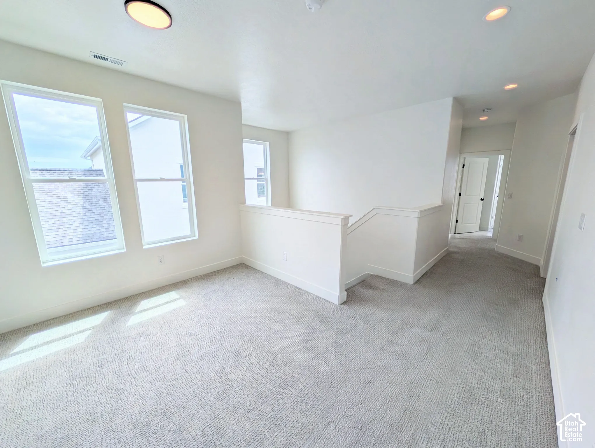 Upper-level space with plush carpeting, multiple windows, a staircase with a white railing, and recessed ceiling lighting
